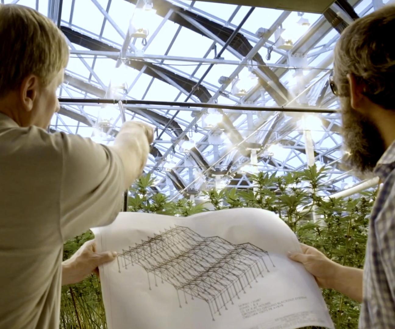 two men standing inside a greenhouse, holding a blueprint-looking sketch of a greenhouse